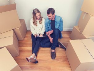 Young couple sitting in new house with packing boxes. They look like they have just moved in and they are very happy, smiling and talking. They are using a digital tablet, sitting on the floor in casual clothes. Copy space.